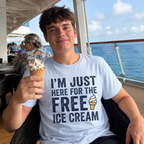 A young man sitting on a cruise ship balcony wearing a light blue graphic t-shirt that reads 'I'M JUST HERE FOR THE FREE ICE CREAM' with a small ice cream cone illustration. He is holding an ice cream cone in his hand, with the ocean and other passengers visible in the background.